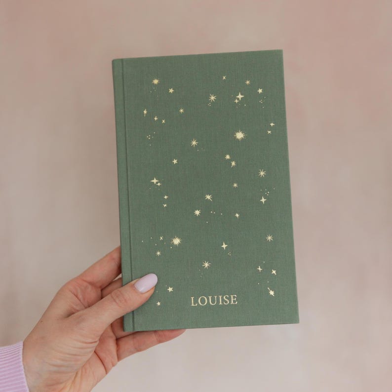 May include: A green hardcover journal with a celestial design of gold stars. The word "LOUISE" is printed in a simple font at the bottom of the cover. The journal is held by a person.