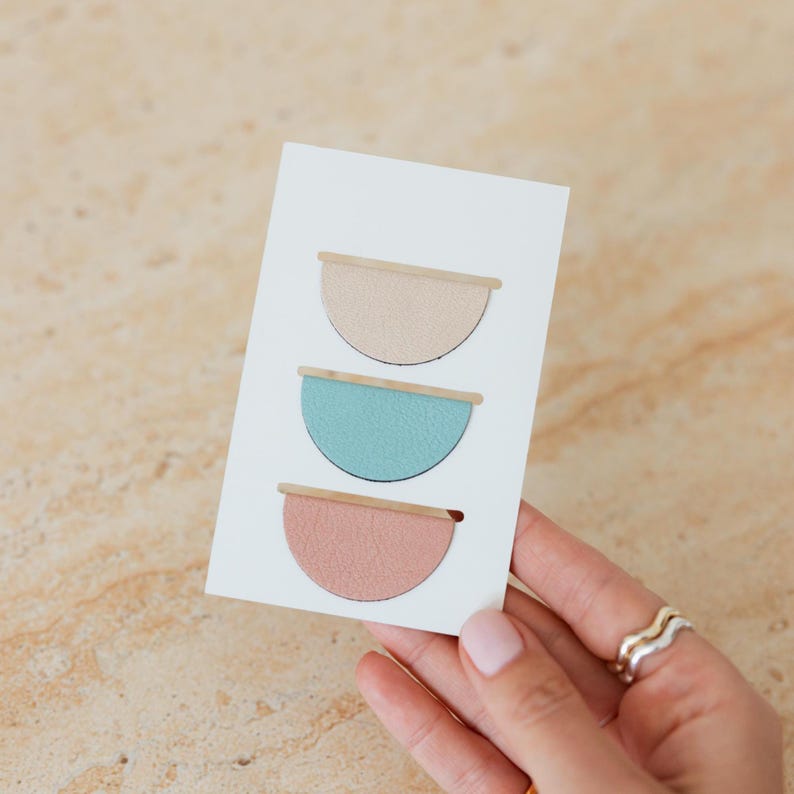 May include: A white card featuring three semi-circular designs in pastel colors: beige, teal, and pink. Each design has a thin, light brown bar across the top. The card is held by a hand with silver rings.