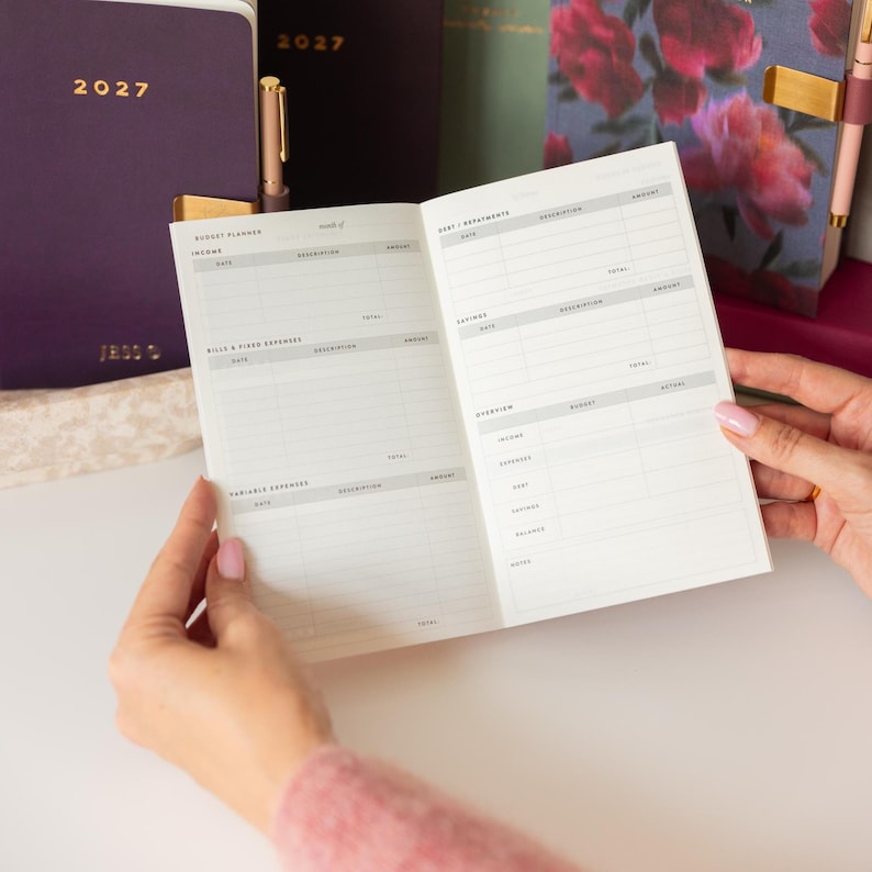 May include: An open budget planner with white pages featuring printed sections for income, bills, fixed expenses, variable expenses, debt, savings, and a monthly overview. The planner has black text on a white background and is held open by two hands.