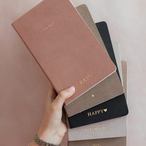 K&ouml;nnte beinhalten: Ein Stapel aus sechs rechteckigen Notizb&uuml;chern in verschiedenen Farben wie Altrosa, Taupe, Schwarz und Hellgrau. Das oberste Notizbuch tr&auml;gt die goldgepr&auml;gten Schriftz&uuml;ge THE DAILY und KATO. Weitere Notizb&uuml;cher zeigen goldene Details wie einen Stern, HAPPY mit einem Herz, den Namen GEORGIE und die Jahreszahl 2026.