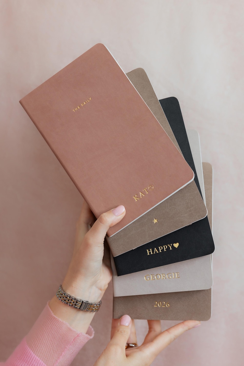 K&ouml;nnte beinhalten: Ein Stapel aus sechs rechteckigen Notizb&uuml;chern in verschiedenen Farben wie Altrosa, Taupe, Schwarz und Hellgrau. Das oberste Notizbuch tr&auml;gt die goldgepr&auml;gten Schriftz&uuml;ge THE DAILY und KATO. Weitere Notizb&uuml;cher zeigen goldene Details wie einen Stern, HAPPY mit einem Herz, den Namen GEORGIE und die Jahreszahl 2026.