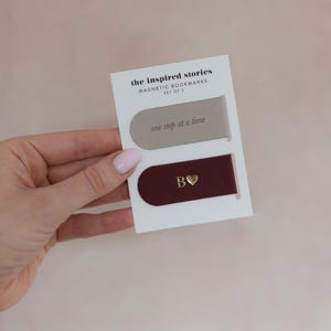 May include: Set of two magnetic bookmarks on a white card. One bookmark is taupe with the text "one step at a time." The other is burgundy with a gold "B" and heart. The card reads "the inspired stories MAGNETIC BOOKMARKS SET OF 2."