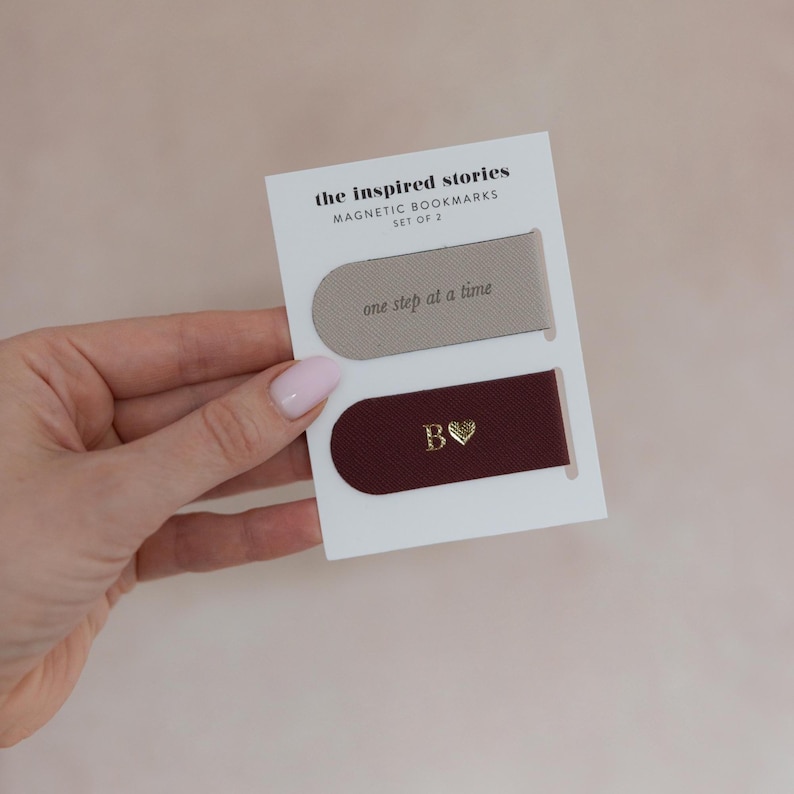 May include: Set of two magnetic bookmarks on a white card. One bookmark is taupe with the text "one step at a time." The other is burgundy with a gold "B" and heart. The card reads "the inspired stories MAGNETIC BOOKMARKS SET OF 2."