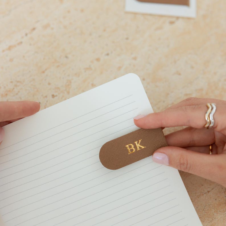 May include: A brown leather bookmark with the gold letters "BK" is placed on the corner of a lined notebook page. The bookmark is held by a person's hand. The background is a light beige color.