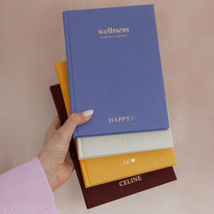 May include: A stack of four hardback journals in blue, cream, yellow, and dark red. The top blue journal features gold text reading wellness EVERYDAY JOURNAL and HAPPY with a smiley face. The other journals have gold initials or names.