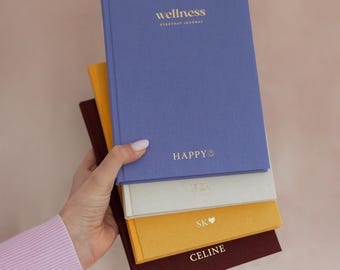 May include: A stack of four hardback journals in blue, cream, yellow, and dark red. The top blue journal features gold text reading wellness EVERYDAY JOURNAL and HAPPY with a smiley face. The other journals have gold initials or names.