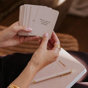 May include: A stack of cream-colored cards with the text "What has surprised you most about life or relationships?" held over a pink notebook and pen. The notebook has the word "twenty" printed on the cover.