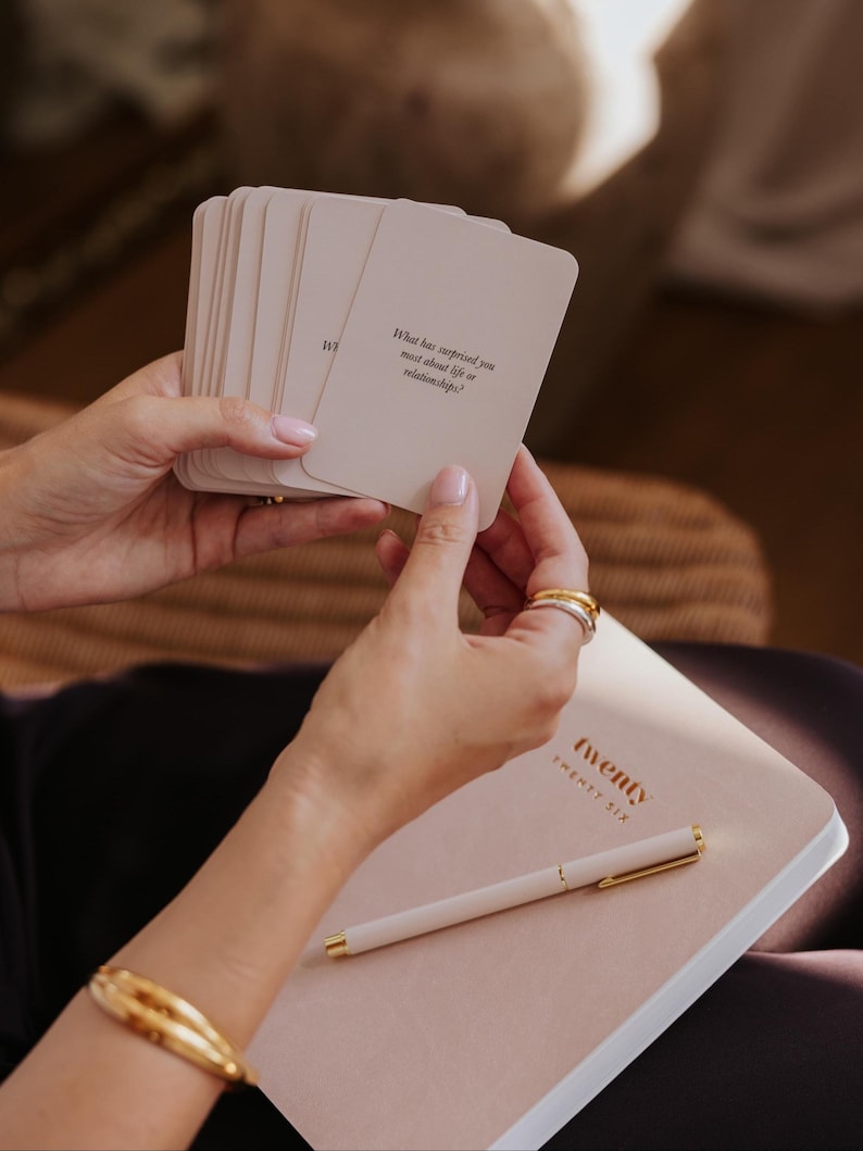 May include: A stack of cream-colored cards with the text "What has surprised you most about life or relationships?" held over a pink notebook and pen. The notebook has the word "twenty" printed on the cover.