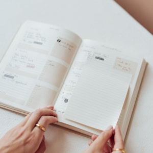 May include: An open planner with lined pages and a blank sticky note. The planner has a beige cover and is open to a page with handwritten notes and a few small stickers.