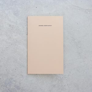 May include: A rectangular notebook with a solid light beige cover. The text "monthly habit tracker" is printed in small, black, sans-serif font near the top center of the cover. The notebook has a clean, simple design with a smooth, matte finish.