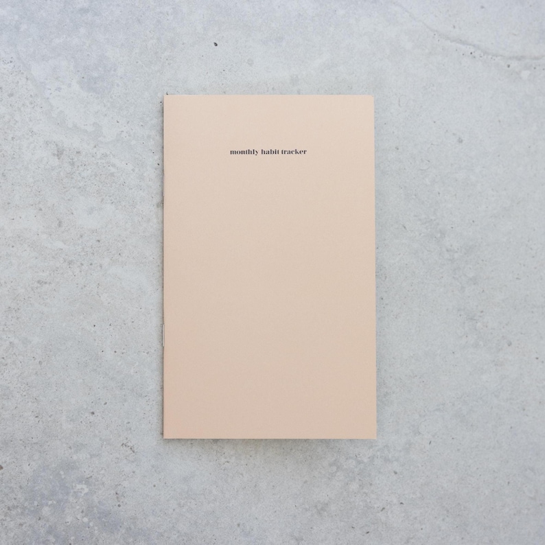May include: A rectangular notebook with a solid light beige cover. The text "monthly habit tracker" is printed in small, black, sans-serif font near the top center of the cover. The notebook has a clean, simple design with a smooth, matte finish.