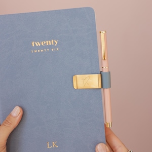 May include: A blue notebook with the word "twenty" in gold lettering. A gold pen clip holds a pink pen. The notebook has the initials "LK" in gold. The background is a solid pink color.
