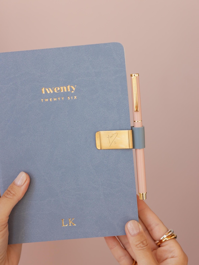 May include: A blue notebook with the word "twenty" in gold lettering. A gold pen clip holds a pink pen. The notebook has the initials "LK" in gold. The background is a solid pink color.