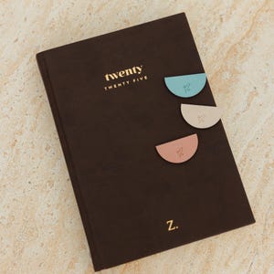 May include: A dark brown journal with the word "twenty" in gold lettering. Three pastel-colored, semi-circular page markers are attached to the journal. The letter "Z" is at the bottom.