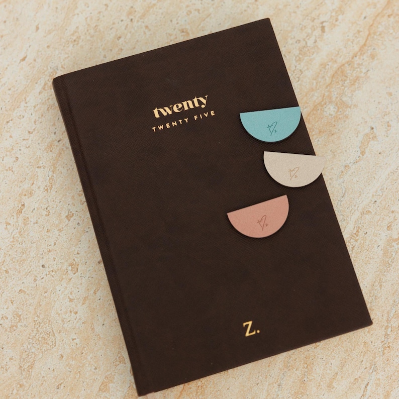 May include: A dark brown journal with the word "twenty" in gold lettering. Three pastel-colored, semi-circular page markers are attached to the journal. The letter "Z" is at the bottom.