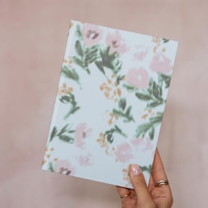 Puede incluir: Un cuaderno rectangular con una cubierta de color azul claro que presenta un patrón floral de enfoque suave en tonos rosa pálido, verde apagado y amarillo dorado. El diseño consiste en flores dispersas y tallos con hojas en la superficie.