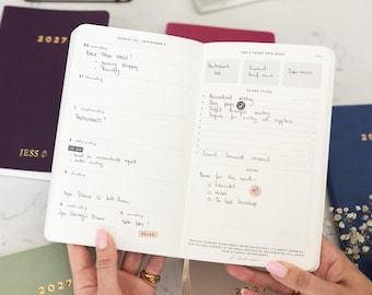 May include: An open weekly planner with white pages featuring black printed text and handwritten notes. The left page shows a daily schedule from Monday to Sunday, while the right page includes sections for top tasks, other tasks, and notes.