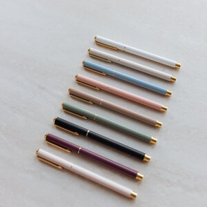 May include: A collection of nine slim ballpoint pens arranged in a diagonal row. Each pen features a solid matte-colored barrel in shades of white, beige, blue, pink, tan, sage green, black, plum, and cream, accented with gold-toned clips and tips.