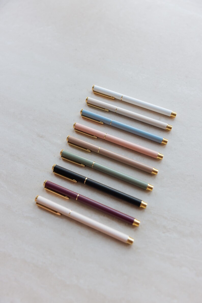 May include: A collection of nine slim ballpoint pens arranged in a diagonal row. Each pen features a solid matte-colored barrel in shades of white, beige, blue, pink, tan, sage green, black, plum, and cream, accented with gold-toned clips and tips.