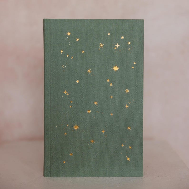 May include: A hardcover journal with a green cover adorned with gold foil star designs. The journal's cover has a textured appearance, and the stars vary in size and arrangement, creating a celestial theme.