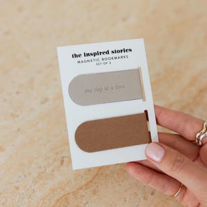 May include: A set of two magnetic bookmarks from "the inspired stories." One bookmark is taupe, the other is brown. The text "one step at a time" is printed on the taupe bookmark. The bookmarks are on a white card.