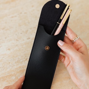 K&ouml;nnte beinhalten: Ein schwarzes Stifteetui mit abgerundeter Klappe und einem goldfarbenen Druckknopfverschluss. Im Etui befinden sich zwei Stifte, einer in Schwarz und einer in Hellrosa, beide mit goldfarbenen Clips und Akzenten. Das Etui hat eine glatte, matte Oberfl&auml;che.