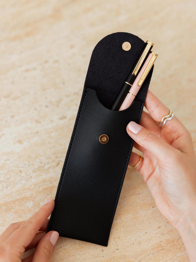 K&ouml;nnte beinhalten: Ein schwarzes Stifteetui mit abgerundeter Klappe und einem goldfarbenen Druckknopfverschluss. Im Etui befinden sich zwei Stifte, einer in Schwarz und einer in Hellrosa, beide mit goldfarbenen Clips und Akzenten. Das Etui hat eine glatte, matte Oberfl&auml;che.