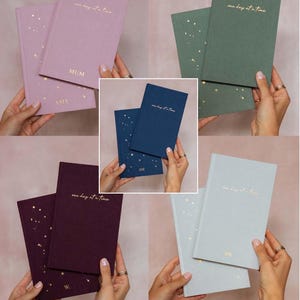 May include: A collection of hardcover journals in various colors, including lavender, green, navy, burgundy, and light gray. Each journal features the phrase "one day at a time" in gold lettering and includes gold star accents. Some journals are personalized.