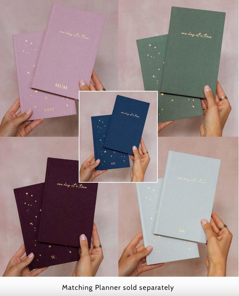 May include: A collection of hardcover journals in various colors, including lavender, green, navy, burgundy, and light gray. Each journal features the phrase "one day at a time" in gold lettering and includes gold star accents. Some journals are personalized.