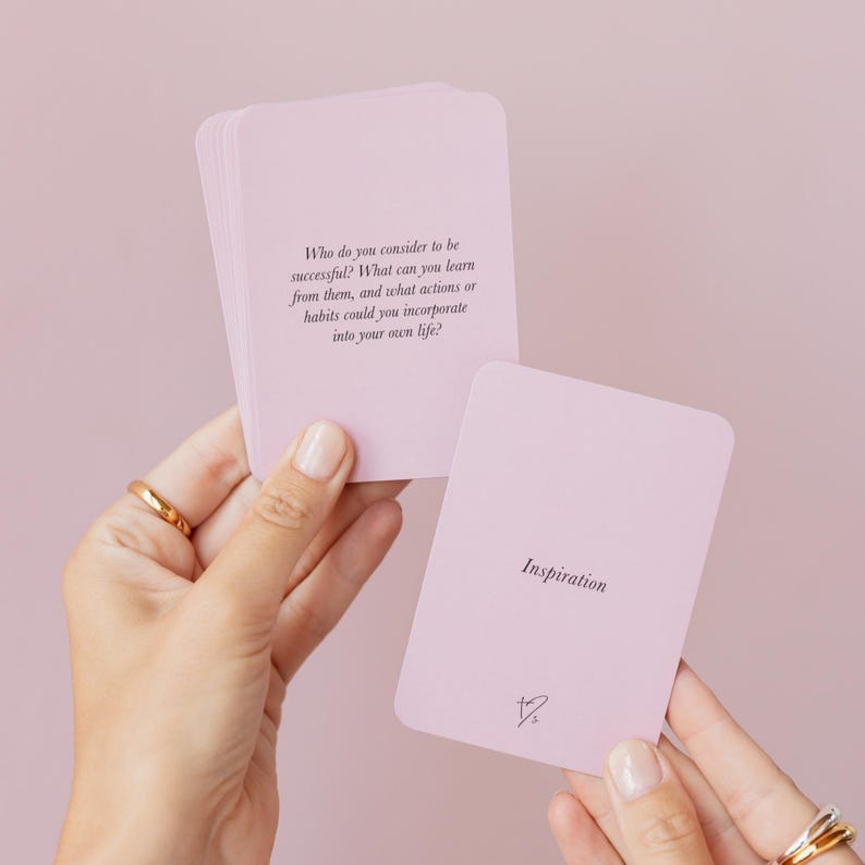 May include: A stack of light pink cards with rounded corners. One card displays the word "Inspiration" and a small symbol. Another card has a question printed on it. The cards are held by hands with gold rings.