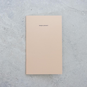 May include: A rectangular budget planner with a solid light beige cover. The words budget planner are printed in a small, black, sans-serif font centered near the top of the front cover. The planner has a clean, flat, and matte appearance.