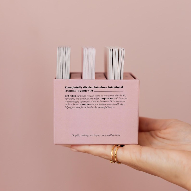 May include: A pink box containing three stacks of white cards. The box has text that reads "Thoughtfully divided into three intentional sections to guide you." The text below describes the cards' purpose: reflection, inspiration, and growth.