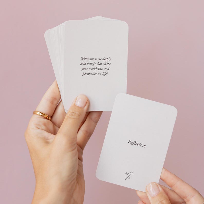 May include: A stack of white cards with rounded corners, held in a person's hands. One card displays the word "Reflection" and a small symbol. Another card has the text: "What are some deeply held beliefs that shape your worldview and perspective on life?"