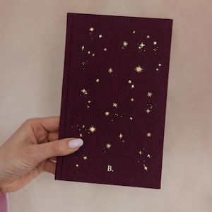 May include: A burgundy journal with a constellation design in gold. The cover features a pattern of stars and celestial shapes. The letter "B" is printed in gold on the lower right corner of the journal.