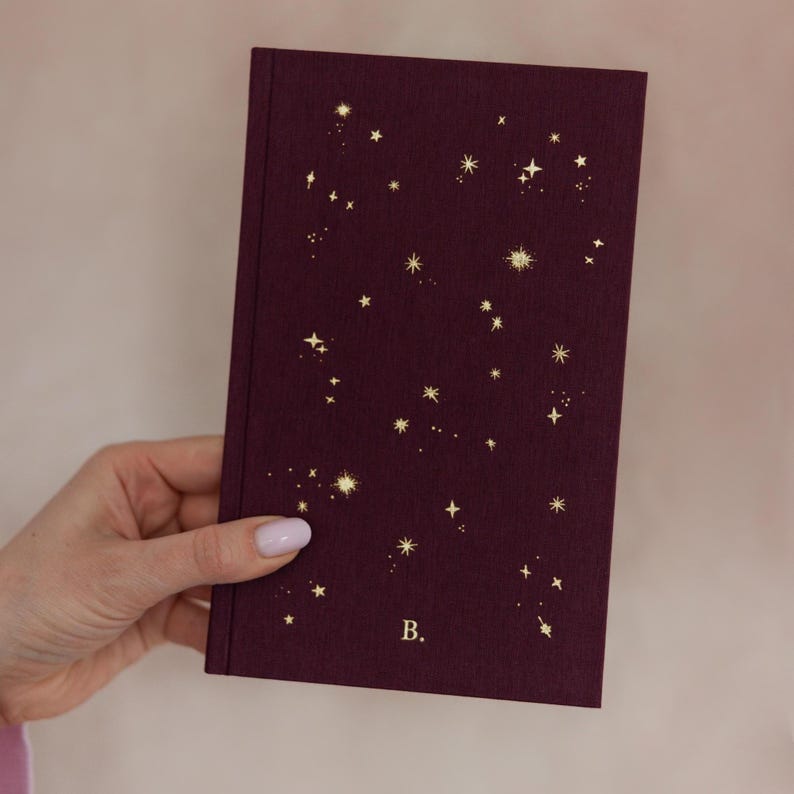 May include: A burgundy journal with a constellation design in gold. The cover features a pattern of stars and celestial shapes. The letter "B" is printed in gold on the lower right corner of the journal.