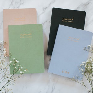Może przedstawiać: Cztery różne kolorowe zeszyty z napisem "inspired" i "bullet journal" napisanym złotymi literami na przedniej stronie. Zeszyty leżą na białym marmurowym blacie. Jeden zeszyt jest jasnoniebieski z napisem "LOVE" napisanym złotymi literami na przedniej stronie.