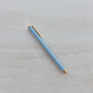 May include: A light blue ballpoint pen with a gold-colored clip and gold-colored accents at the tip and center. The pen has a smooth, matte finish and a slender, cylindrical body, positioned diagonally across the frame with the clip facing upward.