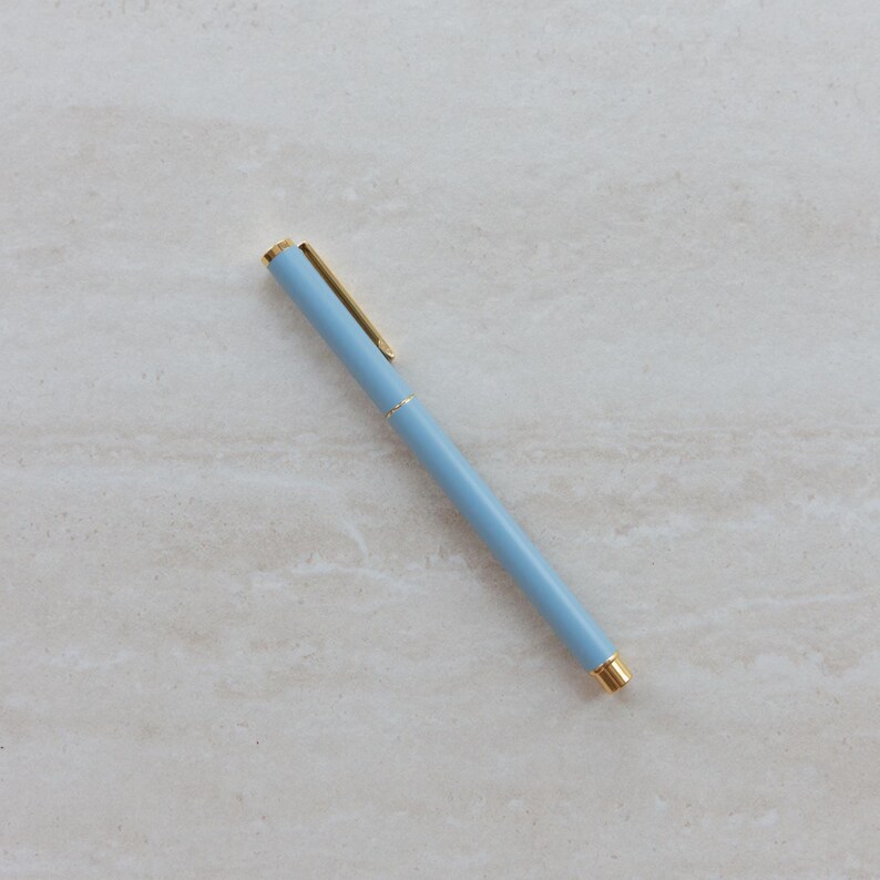 May include: A light blue ballpoint pen with a gold-colored clip and gold-colored accents at the tip and center. The pen has a smooth, matte finish and a slender, cylindrical body, positioned diagonally across the frame with the clip facing upward.