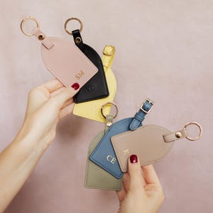 Könnte beinhalten: Eine Sammlung von sechs bogenförmigen Kofferanhängern in verschiedenen Farben wie Hellrosa, Schwarz, Gelb, Olivgrün, Blau und Beige. Jeder Anhänger hat einen goldfarbenen Metallring, einen passenden Schnallenriemen und goldgeprägte Initialen: SM, VO, TV, DG, CE und K.