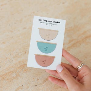 May include: A set of three magnetic bookmarks in a card. Each bookmark is a half-circle with a wooden top and a small heart design. The colors are beige, teal, and coral. The card reads "the inspired stories MAGNETIC BOOKMARKS SET OF 3."
