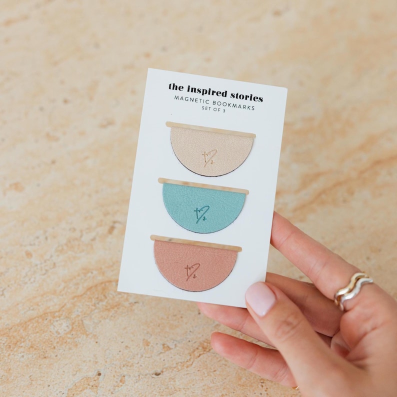 May include: A set of three magnetic bookmarks in a card. Each bookmark is a half-circle with a wooden top and a small heart design. The colors are beige, teal, and coral. The card reads "the inspired stories MAGNETIC BOOKMARKS SET OF 3."