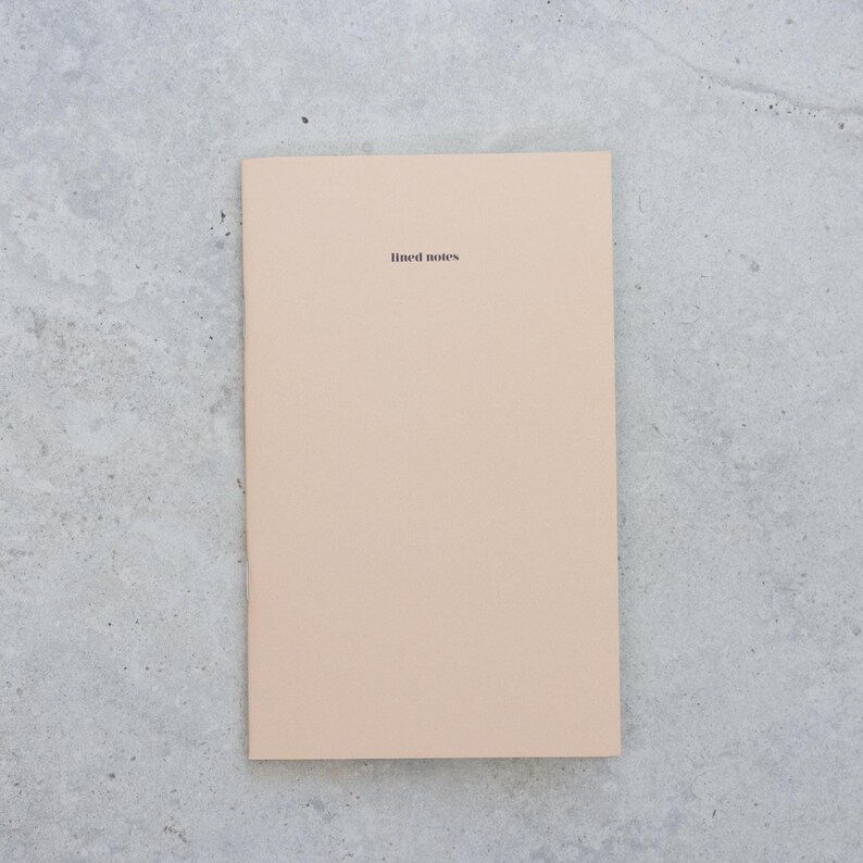 May include: A rectangular notebook with a solid light beige cover. The words "lined notes" are printed in a small, black, sans-serif font centered near the top of the front cover. The notebook has a clean, simple design with a smooth, matte finish.