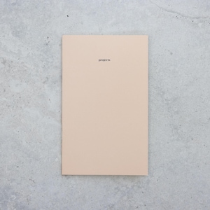 May include: A rectangular notebook with a solid light beige cover. The word projects is printed in small, black, lowercase sans-serif letters near the top center of the front cover. The notebook has a clean, flat, and uniform appearance.
