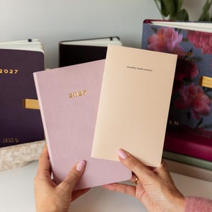May include: A light purple 2027 planner and a beige monthly habit tracker booklet held together. In the background, a dark purple 2027 planner and a floral-patterned planner are visible. The planners feature gold text and clean, simple covers.