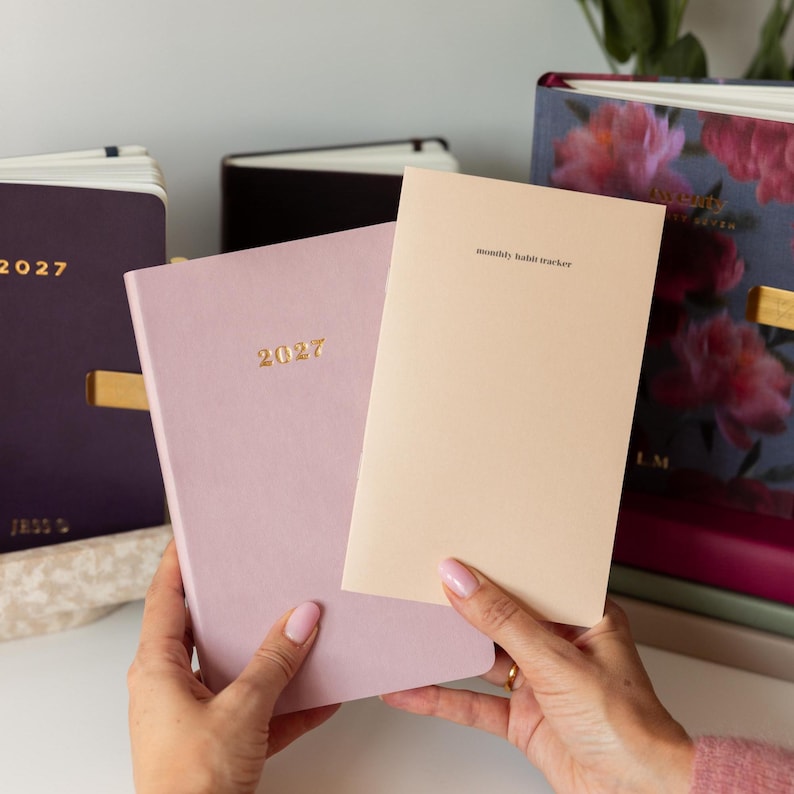 May include: A light purple 2027 planner and a beige monthly habit tracker booklet held together. In the background, a dark purple 2027 planner and a floral-patterned planner are visible. The planners feature gold text and clean, simple covers.