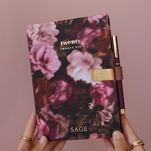May include: A floral print journal with the words "twenty twenty six" and "SAGE" in gold lettering. The journal has a gold clasp and a matching burgundy pen. The background is a solid pink color.