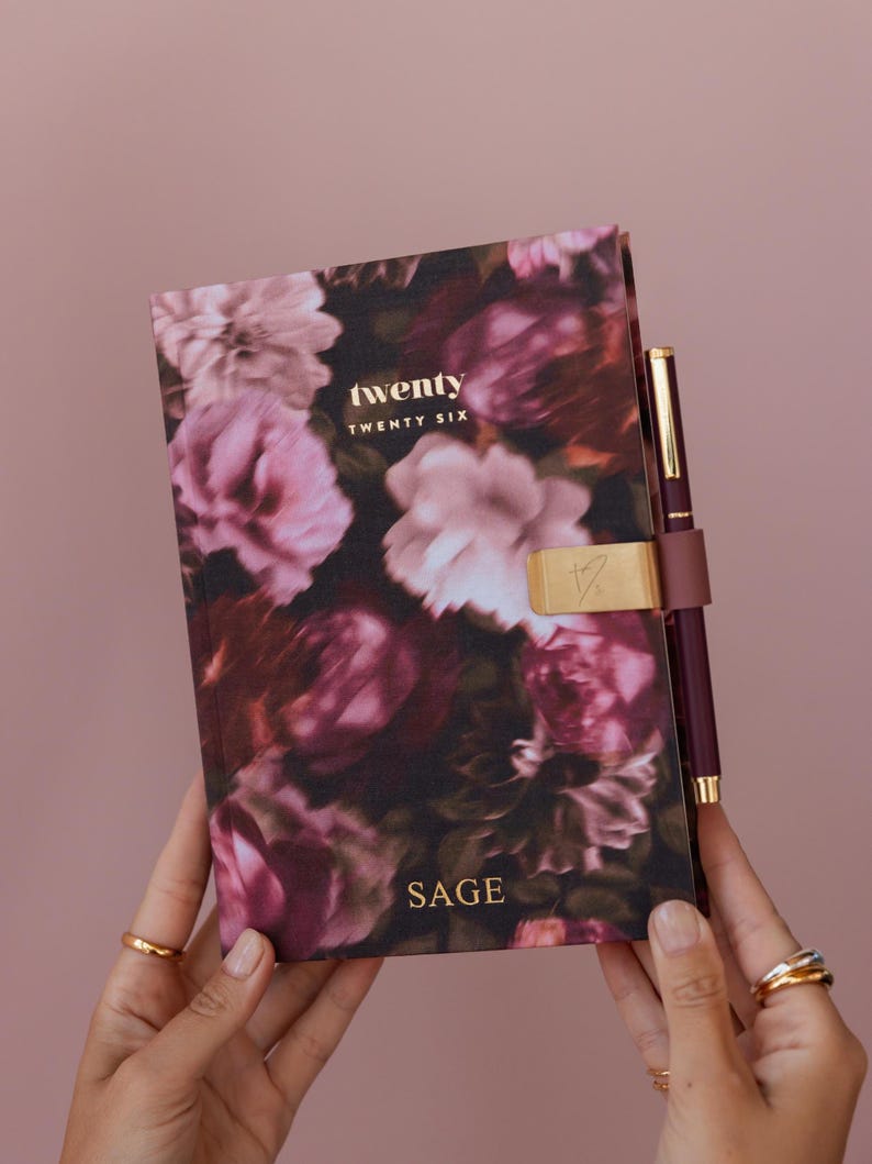 May include: A floral print journal with the words "twenty twenty six" and "SAGE" in gold lettering. The journal has a gold clasp and a matching burgundy pen. The background is a solid pink color.