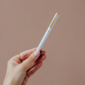 May include: A slim, light blue ballpoint pen featuring a polished gold-tone clip, a matching gold-tone band at the center, and a gold-tone cap top. The pen has a smooth, matte finish and a cylindrical body, presenting a clean and simple design.