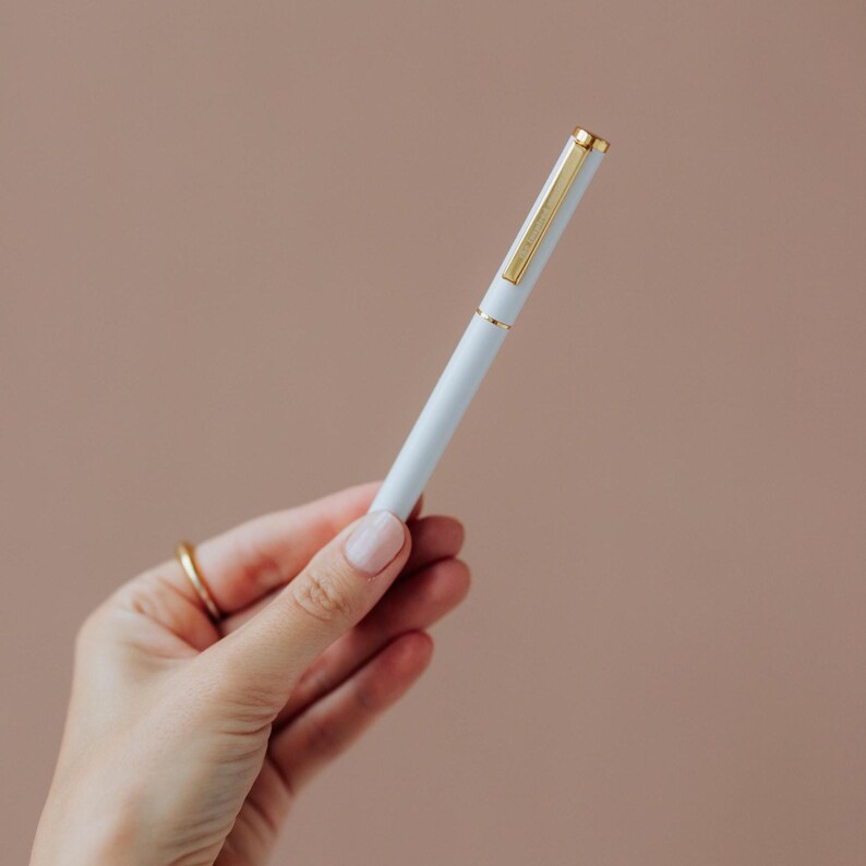 May include: A slim, light blue ballpoint pen featuring a polished gold-tone clip, a matching gold-tone band at the center, and a gold-tone cap top. The pen has a smooth, matte finish and a cylindrical body, presenting a clean and simple design.