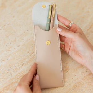 K&ouml;nnte beinhalten: Ein beiges Stifteetui mit goldenem Druckknopfverschluss enth&auml;lt zwei schlanke Stifte, einen in Salbeigr&uuml;n und einen in hellem Beige, beide mit goldenen Clips und Akzenten. Das Etui hat eine geschwungene Klappe und sichtbare Seitenn&auml;hte.
